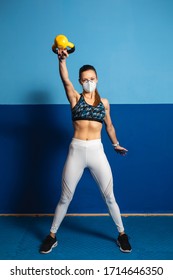 Young fit woman with n96 face mask  doing shoulder exersice at the gym with kettlebells. Fitness strength workout under coronavirus health crisis.