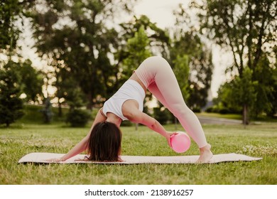 young fit woman pink sportswear and rubber rubber pilates ball does exercises and stands in down dog pose in park in summer. 