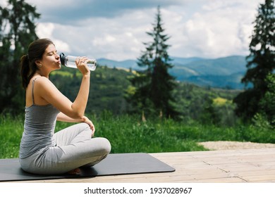 Young fit woman sitting on yoga mat and drinking water after training on background of sunny mountains hills. Outdoor workout. Healthy lifestyle. Sporty Female relaxing on mat among trees - Powered by Shutterstock