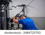 young fitness athlete, seen from behind, is performing a triceps exercise using a high pulley machine in the gym as part of his strength training routine