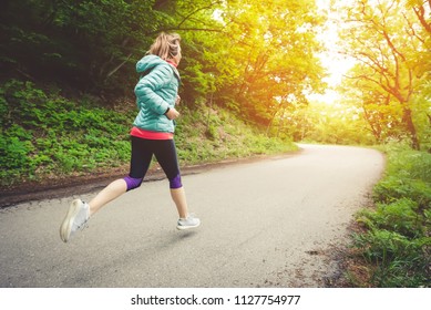 Young Fitness Blonde Woman In Headphones Running At Morning Caucasian Forest Trail In Sun Light. Side View From Behind