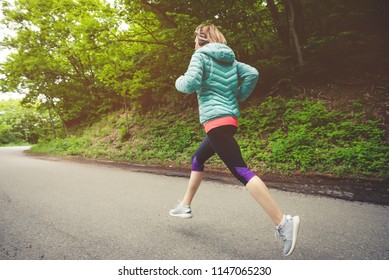 Young Fitness Blonde Woman In Headphones Running At Morning Caucasian Forest Trail In Sun Light. Side View From Behind