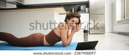 Young fitness instructor, woman does yoga, looks at tablet, watches online video pilates class, meditation training, listens in wireless headphones, lying on rubber mat, gym indoors at home.
