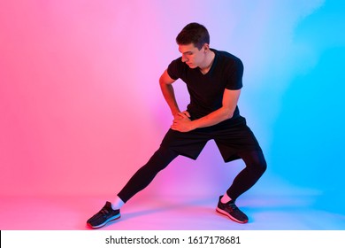 Young Fitness Man With Headphones Doing Stretching Exercises On Background With Red And Blue Neon Light