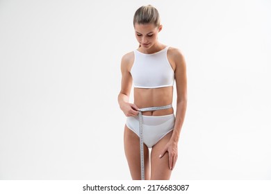 Young Fitness Model Measuring Waist Line Standing On White Background In Sport Outfit, Looking Down On Scale. Flat Stomach. Toned Body, Abs, Keep Fit