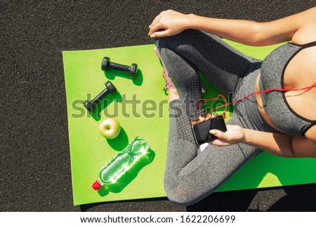 Young fitness sportswoman sitting and using smartphone in gym. Girl on yoga mat with botte of water, dumbbell and ealthy apple listens to music. Sports and healthy concept. top view.