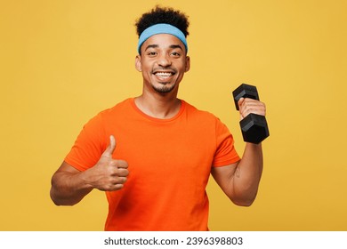 Young fitness trainer instructor sporty man sportsman wear orange t-shirt hold in hand dumbbell show thumb up spend time in home gym isolated on plain yellow background. Workout sport fit abs concept - Powered by Shutterstock