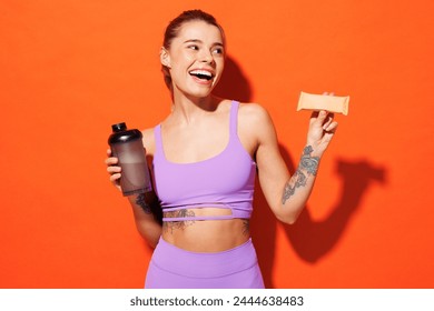 Young fitness trainer instructor sporty woman sportsman wear purple top clothes in home gym eat protein bar hold cocktail look aside isolated on plain orange background Workout sport fit abs concept - Powered by Shutterstock