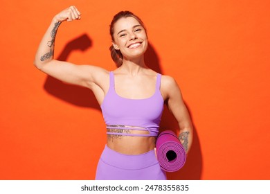 Young fitness trainer instructor sporty woman sportsman wear purple top clothes spend time in home gym hold yoga mat show hand biceps isolated on plain orange background. Workout sport fit abs concept - Powered by Shutterstock