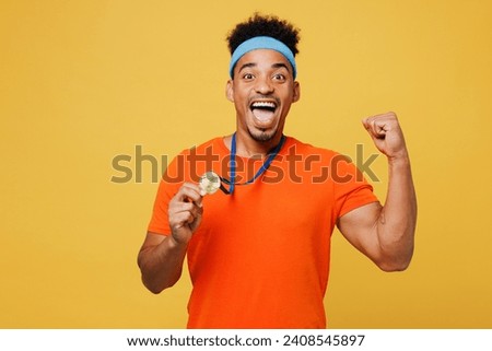 Young fitness trainer instructor sporty man sportsman wear orange t-shirt show golden medal award do winner gesture spend time in gym isolated on plain yellow background. Workout sport fit abs concept