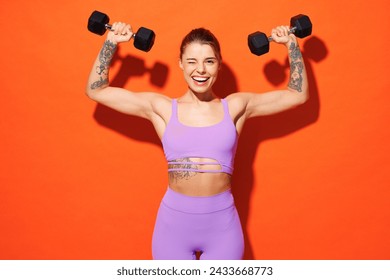 Young fitness trainer instructor woman sportsman wear purple top clothes spend time in home gym hold dumbbells raise up hands wink isolated on plain orange background. Workout sport fit abs concept - Powered by Shutterstock