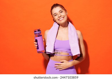 Young fitness trainer sporty woman sportsman wear purple top clothes spend time in home gym hold water bottle towel put hand on belly isolated on plain orange background. Workout sport fit abs concept Stock fotografie