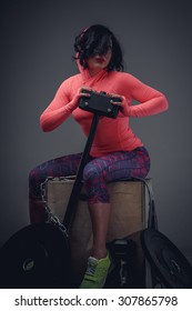 Young Fitness Woman With Black Hair In Pink Shirt And Violet Pants Sitting On Wooden Box And Holding Big Black Hummer.