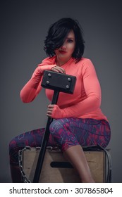 Young Fitness Woman With Black Hair In Pink Shirt And Violet Pants Sitting On Wooden Box And Holding Big Black Hummer.