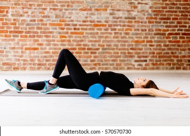 Young fitness woman in black sportswear doing stretching exercise with foam roller  in modern loft interior at gym. Sport, fitness, lifestyle and people concept