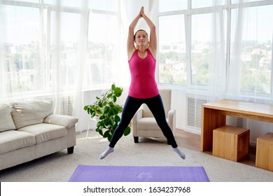 Young fitness woman doing jumping jacks or star jump exercise at home, copy space. Girl working out, full length portrait. Healthy lifestyle concept