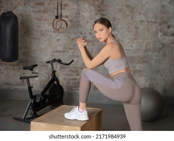 Young Fitness Woman In Fitwear Holding Hands Clasped, Looking At Camera And Working Out On A Brick Wall, Exercise Bike, Punching Bag And Fitness Ball Background.

Sport And Healthy Lifestyle Concept.