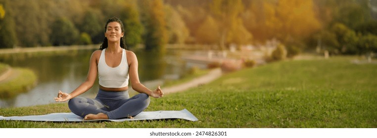 Young fitness woman in fitwear practices morning yoga sitting in lotus position, meditating on mat in nature near lake in green park. Lady enjoys calmness during workout meditation, copy space - Powered by Shutterstock