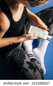 Young Fitness Woman On Yoga Mat Holding Phone Online Application Track Menstrual Cycle. Sport At PMS Critical Days Concept. Female Health Care, Gynecological Concept. Ovulation Calendar On Mobile App