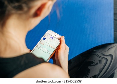 Young Fitness Woman On Yoga Mat Holding Phone Online Application Track Menstrual Cycle. Sport At PMS Critical Days Concept. Female Health Care, Gynecological Concept. Ovulation Calendar On Mobile App