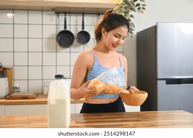 Young fitness woman pouring cornflakes into a wooden bowl, preparing breakfast on kitchen counter - Powered by Shutterstock