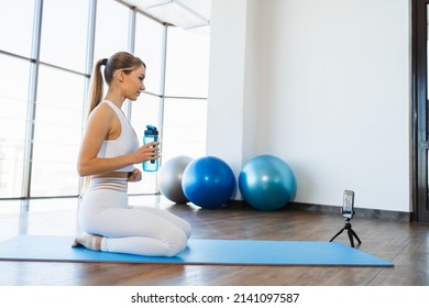 Young Fitness Woman Sitting On Yoga Mat With Bottle Of Water And Live Streaming Workout