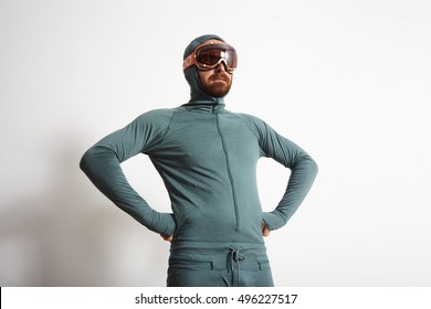 Young Fitted Bearded Male Athlete In Baselayer Thermal Suite Wears Snowboarding Googles, Proudly Stands In Front Of White Wall