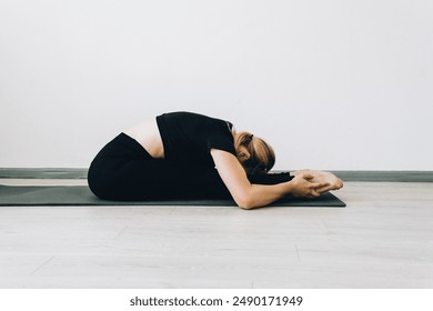 A young flexible girl does stretching and does yoga on a sports mat. Shooting from the side. paschimottanasana pose, training. Gymnastics and yoga classes in the studio - Powered by Shutterstock