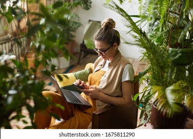 Young florist in eyeglasses sitting on armchair with laptop computer on her knees and using mobile phone in the room with plants - Powered by Shutterstock