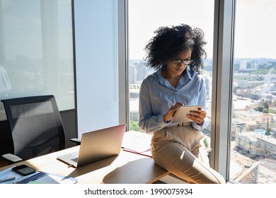 Young focused African American businesswoman sitting on desk holding using tablet computer device in modern office in high floor building with panoramic urban capital city view.