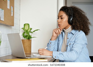 Young Focused African Girl Woman Lady Student Sit Table Look At Laptop Screen Wear Headphones At Home Office Study, Elearning Online Course Conference Zoom Hangout Call Remote Education Work Concept.