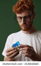 Young focused caucasian clever red hair guy playing rubik's cube in hands. Concept of human of generation Z. Handsome bearded male person in glasses. Isolated on dark green background in studio