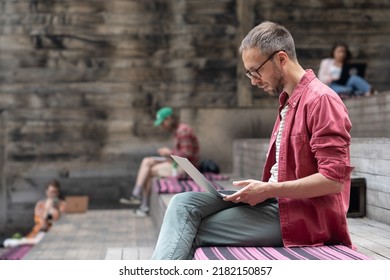 Young focused creative man freelance web designer using laptop while working in outdoor space, student guy sitting on wooden bench with computer, surfing internet, studying outside. Selective focus