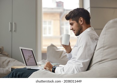 Young focused european businessman drinking tea or coffee from cup and watching business graph on laptop. Concept of modern successful man. Bearded stylish guy wearing suit. Office interior. Daytime