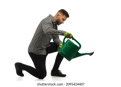 Young focused man in lumberjack shirt is kneeling and watering by using a watering can. Side view. Full length studio shot on white background. - Powered by Shutterstock