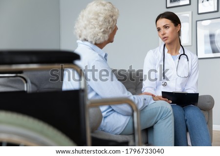 Young focused nurse visiting mature middle aged disabled patient for rehabilitation therapy at home. Skilled attentive millennial physiotherapist consulting senior old woman, healthcare concept.