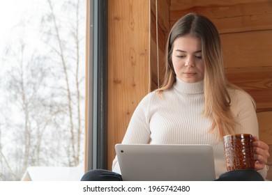 Young focused woman holding a coffee mug sitting next to the window cross-legged. Working on her laptop in a peaceful room. Winter season. Snow-covered trees can be seen outside through the window. 