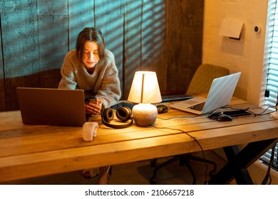 Young focused woman using smart phone while sitting at cozy home office during the nighttime. Concept of online work from home. Idea of cozy and style workplace