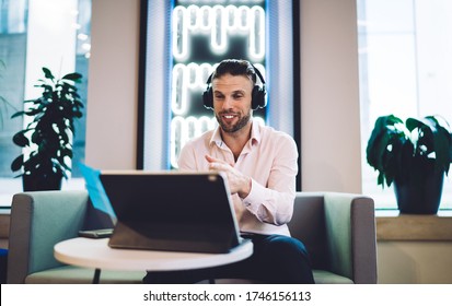 Young Formally Dressed Male In Headphones Using Tablet Computer For Online Meeting While Sitting At Round Table In Open Office Space