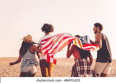 Young And Free. Rear View Of Four Young People Carrying American Flag While Running Outdoors