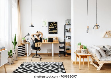 Young freelance woman working remotely at designer home in cozy white office room with pendant lamps, white wall, big window, comfy couch and aloe plant standing on wooden retro table - Powered by Shutterstock