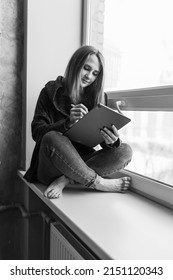 Young Freelancer Girl On The Windowsill With A Tablet. Freelance Artist Woman Working From Home. Home Office, Freelance.