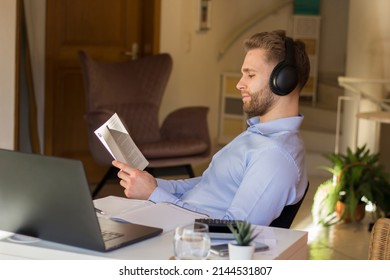 Young Freelancer Reading A Book While Taking A Break In His Home Office. Wearing A Noise Canceling Headphone While Reading.