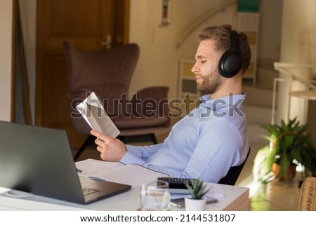 Young freelancer reading a book while taking a break in his home office. Wearing a noise canceling headphone while reading.