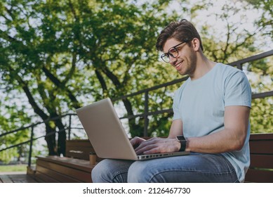 Young freelancer smiling happy student man in blue t-shirt eyeglasses sit on bench work with laptop pc computer rest relax in sunshine spring green city park outdoors on nature. Urban leisure concept