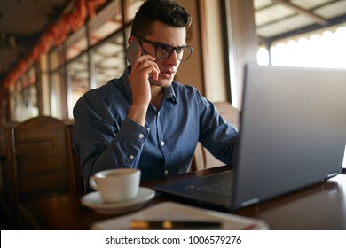 Young freelancer working with laptop and talking on cellphone with client in office. Handsome caucasian businessman in glasses conducts negotiations via phone call. Multitasking business concept.