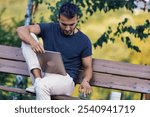 Young freelancer working remotely with his laptop, sitting on a bench in a park, enjoying the fresh air and nature while staying connected and productive