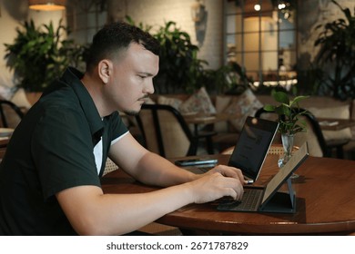 Young freelancer writing notes in notebook while working on laptop at cozy cafe table. Concept of productivity, business planning, studying, freelance career, and digital entrepreneurship.