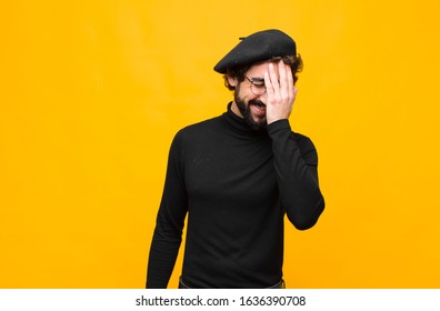 Young French Artist Man Laughing And Slapping Forehead Like Saying D’oh! I Forgot Or That Was A Stupid Mistake Against Orange Wall