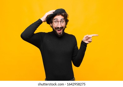 Young French Artist Man Laughing, Looking Happy, Positive And Surprised, Realizing A Great Idea Pointing To Lateral Copy Space Against Orange Wall